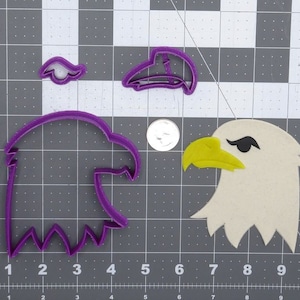 May include: Purple plastic cookie cutters in the shape of an eagle's head, beak, and eye. The cutters are on a gray grid background.