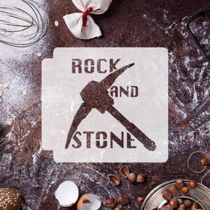 May include: White stencil with the text "Rock and Stone" and a pickaxe graphic. The stencil is on a brown surface with flour scattered around it.