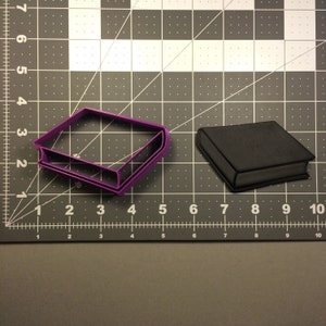 Puede incluir: Dos cortadores de galletas con forma de libro, uno morado y otro negro, sobre una superficie de rejilla gris.