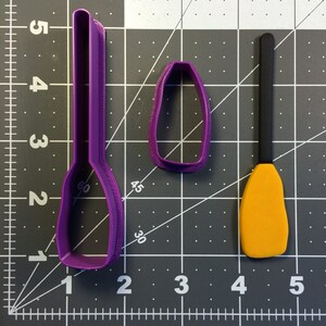 Può includere: Tre stampini per biscotti in plastica viola, uno a forma di pagaia, uno a forma di canoa e uno a forma di ovale piccolo. Un biscotto di pasta di zucchero giallo è su un tappetino da taglio bianco e nero.