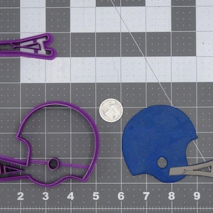 Pode incluir: Um cortador de biscoitos de capacete de futebol roxo, um recorte de capacete de futebol azul e um cortador de biscoitos de máscara facial roxa são exibidos em uma grade cinza. Uma moeda está ao lado dos recortes do capacete. Os cortadores de biscoitos são para assar.