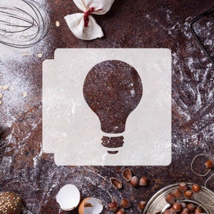 Può includere: Uno stencil bianco con un disegno di lampadina su una superficie marrone ricoperta di farina bianca. Lo stencil è circondato da vari ingredienti da forno, tra cui noci, gusci d'uovo e una frusta.