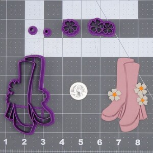 May include: Purple plastic cookie cutters in the shape of a pair of platform boots and two flowers. The cutters are on a gray grid background.