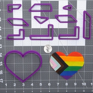 May include: A set of purple plastic cookie cutters in various shapes, including hearts, squares, and rectangles. A rainbow heart-shaped cookie is also shown on a gray grid.