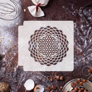 May include: White plastic stencil with a flower of life design. The stencil is on a brown surface with flour scattered around it.