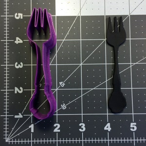 May include: Purple plastic fork cookie cutter with a black plastic fork next to it on a gridded surface.