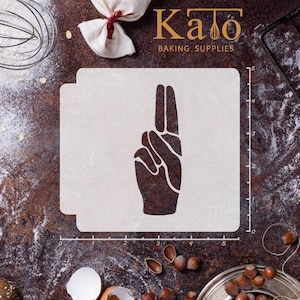 May include: A white plastic stencil with a hand making the peace sign. The stencil is 5 inches wide and 5 inches tall. The stencil is on a brown surface with flour and nuts.