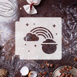 May include: White stencil with a rainbow and a pot of gold at the end of the rainbow. The stencil is on a brown surface with flour scattered around it.