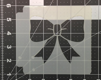 May include: A white plastic stencil of a black bow. The stencil is on a black and white grid background.
