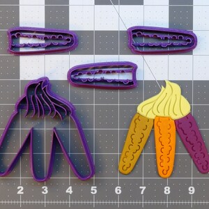 May include: A set of purple plastic cookie cutters in the shape of corn on the cob. The cutters are on a grey grid background.