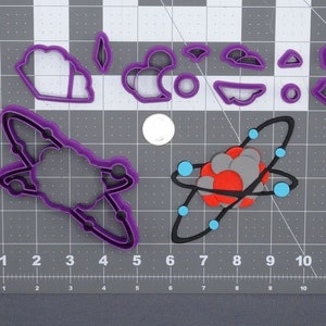 May include: A set of purple plastic cookie cutters in the shape of an atom, a rocket ship, and clouds. The atom cookie cutter has a red and gray center with blue circles around it. The cookie cutters are on a gray grid surface.