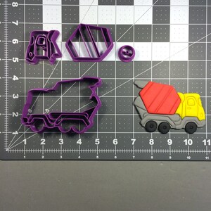Puede incluir: Un juego de tres cortadores de galletas de plástico morado con forma de camión mezclador de cemento. El camión mezclador de cemento se muestra con un diseño rojo y gris. El juego también incluye un camión más pequeño y una forma redonda.