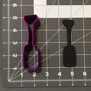 Può includere: Tagliapasta in plastica viola a forma di sbattitore. Il tagliapasta è mostrato accanto a un tagliapasta in plastica nera della stessa forma.