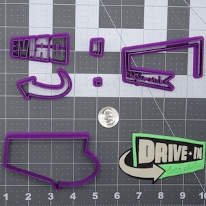 Puede incluir: Cortadores de galletas de plástico morado en forma de letrero de cine de autocine, boleto de cine de autocine, flecha de cine de autocine y la palabra "DRIVE" con una flecha apuntando hacia la derecha. También hay un cortador de galletas de plástico morado en forma de estado.