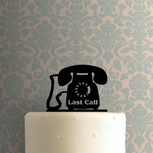 Puede incluir: Un adorno para tarta negro con la silueta de un teléfono antiguo. El teléfono tiene un dial giratorio y las palabras "Last Call" están impresas en la base. El adorno está colocado sobre una tarta blanca con un fondo estampado en azul claro y beige.