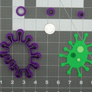 May include: A collection of purple and black cookie cutters in various sizes, shaped like viruses. A green virus-shaped cutout is also present. A coin is included for size comparison. The items are on a gray grid.