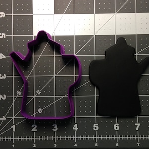 May include: Purple plastic cookie cutter shaped like a teapot. The cutter is on a black and white grid background.