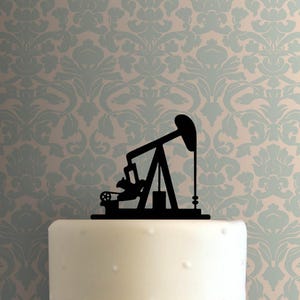 Könnte beinhalten: Ein schwarzer Silhouetten-Kuchenaufsatz mit einem Ölförderpumpen-Motiv. Der Aufsatz befindet sich auf einem weißen Kuchen mit gepunktetem Muster, vor einem Hintergrund aus hellblauer und cremefarbener floraler Tapete.