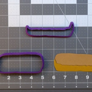 May include: Two purple plastic cookie cutters, one shaped like a canoe and the other shaped like a long rectangle. A brown and yellow cookie cutter shaped like a loaf of bread is in the background.