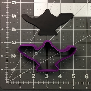 Pode incluir: Um cortador de biscoitos de plástico roxo em forma de lâmpada de gênio. O cortador está em uma superfície quadriculada cinza com linhas brancas.