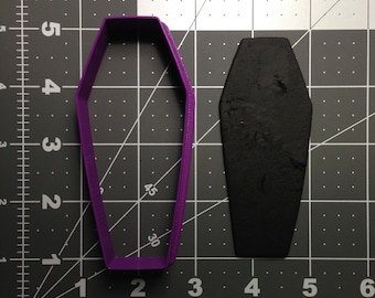 May include: A purple plastic coffin-shaped cookie cutter and a black fondant coffin on a gray grid background.