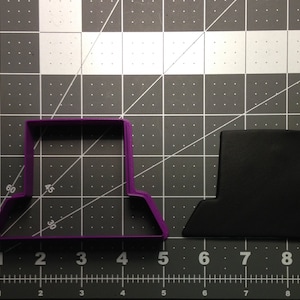 May include: A purple plastic cookie cutter shaped like a hat. The cookie cutter is on a gray grid surface. A black cookie cutter shaped like a hat is to the right of the purple cookie cutter.