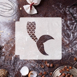 May include: A white plastic stencil with a mermaid tail design. The stencil is on a brown surface with flour scattered around it.