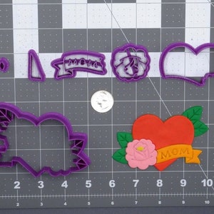 Pode incluir: Um conjunto de cortadores de biscoitos de plástico roxo em forma de coração, rosa, faixa e rosa com espinhos. A faixa diz "MOM".