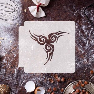 Peut inclure: Un pochoir blanc avec un motif tribal composé de trois volutes et d'une forme d'aile. Le pochoir est sur une surface brune avec de la farine et d'autres ingrédients de cuisson.