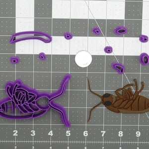 May include: Purple plastic cookie cutters in the shape of a cockroach and various other shapes. A brown plastic cockroach cookie cutter is also shown.