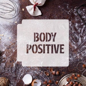 Peut inclure: Un pochoir blanc avec les mots "BODY POSITIVE" imprimés en noir sur un fond brun avec de la farine et d'autres ingrédients de cuisson.