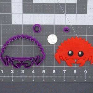 Pode incluir: Um conjunto de três cortadores de biscoitos de plástico roxo e vermelho em forma de caranguejo e concha. Os cortadores estão sobre um fundo de grade cinza.