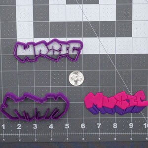 May include: Two purple plastic cookie cutters, one shaped like the word "MACED" in graffiti style and the other shaped like the word "MUSIC" in graffiti style. The "MUSIC" cutter is pink and purple.
