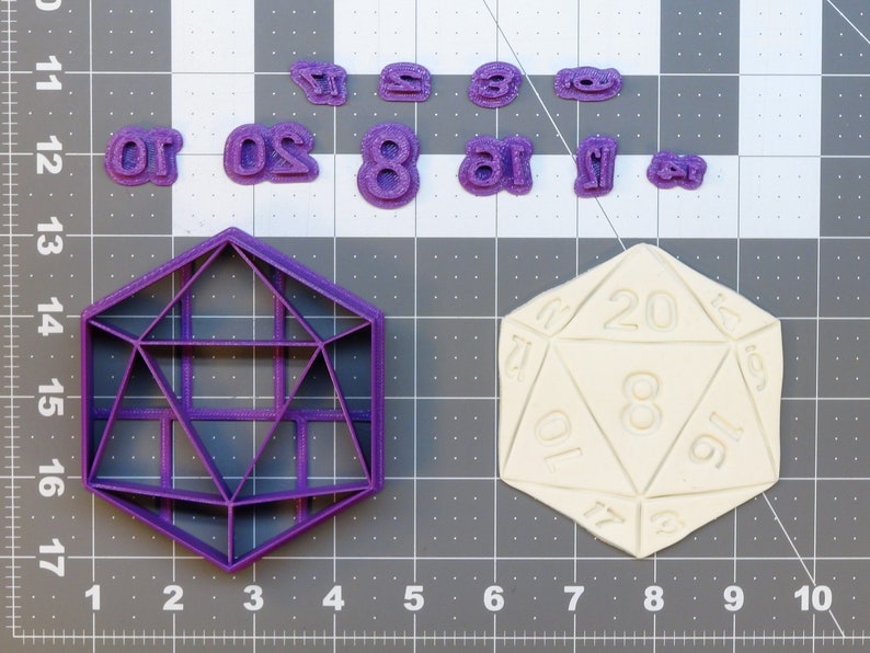 May include: Purple plastic cookie cutters shaped like a 20-sided die and a geometric shape. There are also individual number cookie cutters for 1, 2, 3, 4, 5, 6, 7, 8, 10, 16, 20, and 24. A white plastic 20-sided die is also shown.