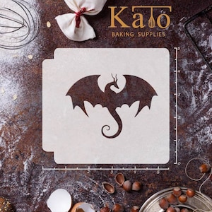 May include: A white plastic stencil with a black silhouette of a dragon with wings spread. The stencil is 5 inches wide and 5 inches tall. The stencil is on a brown wooden surface with flour and nuts.