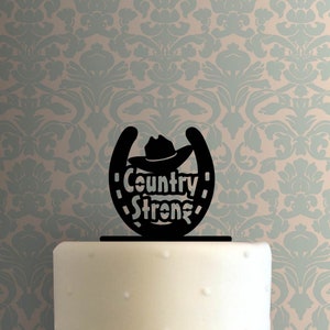 May include: Black silhouette of a horseshoe with a cowboy hat on top and the text "Country Strong" inside. This is a cake topper.