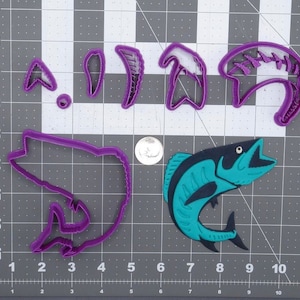 Op de afbeelding: Paarse plastic koekjesvormen in de vorm van een vis, een visvin, een visgraat en een haak. Er is ook een blauwe en groene vis koekjesvorm.