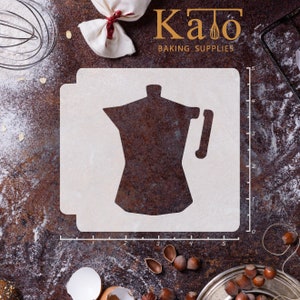May include: White plastic stencil of a coffee pot on a brown background with a ruler marked in inches. The stencil is for baking and decorating.