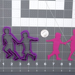 May include: Two purple plastic cookie cutters shaped like children holding hands. The cutters are on a gray grid background.
