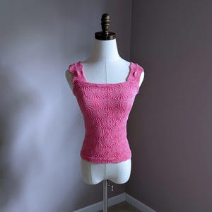 May include: A pink, sleeveless top with a square neckline and a textured, floral-like pattern. The top is displayed on a mannequin, showcasing its fitted silhouette and decorative shoulder straps. The garment is made of a soft, possibly knitted material.