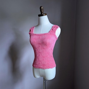 May include: A pink, textured tank top with a square neckline and scalloped edges. The top is displayed on a mannequin. The fabric has a raised, floral-like pattern.