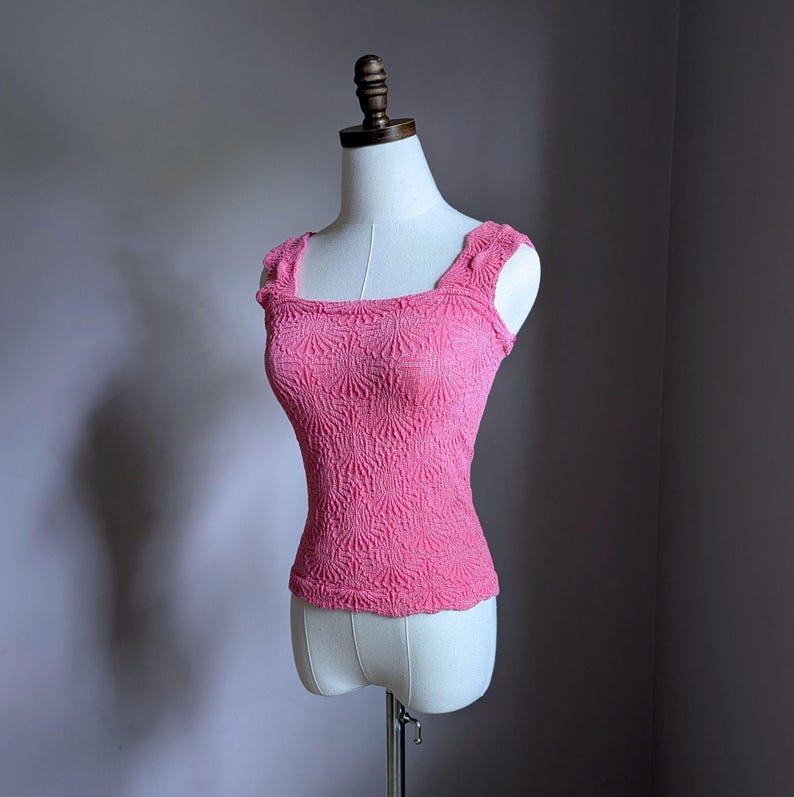 May include: A pink, textured tank top with a square neckline and scalloped edges. The top is displayed on a mannequin. The fabric has a raised, floral-like pattern.