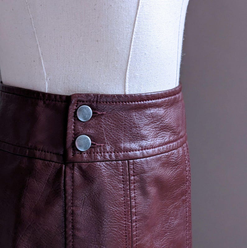 May include: A burgundy faux leather skirt with a high waist and two silver buttons. The skirt has a straight cut and a simple design.