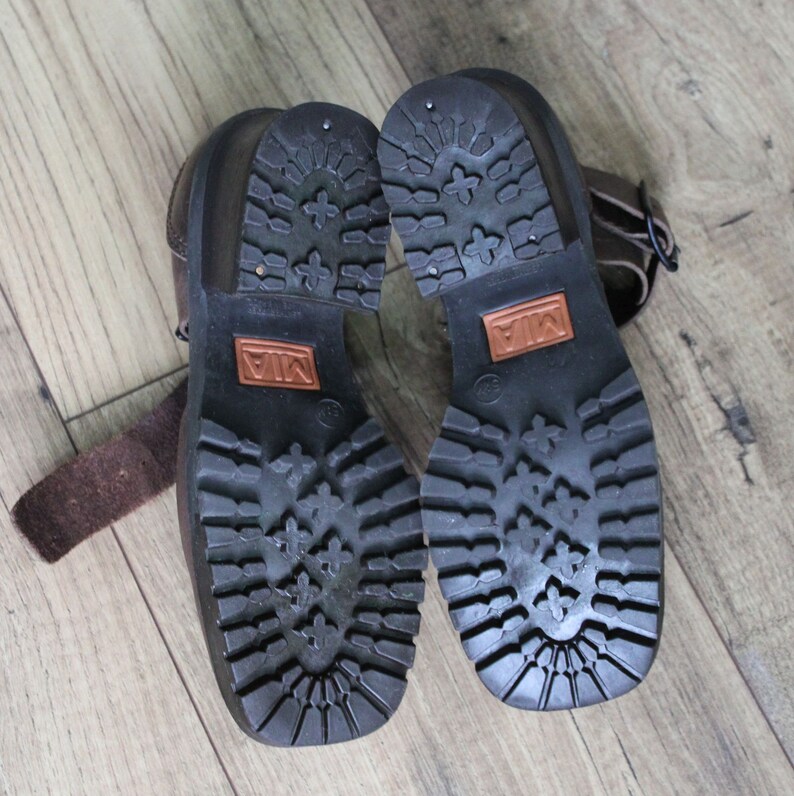 May include: A pair of brown leather ankle boots with a buckle strap. The boots have a black rubber sole with a tread pattern. The brand name "MIA" is printed on the sole.