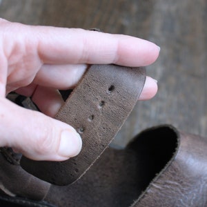 May include: Close-up of a brown leather sandal strap with four holes for adjusting the fit.