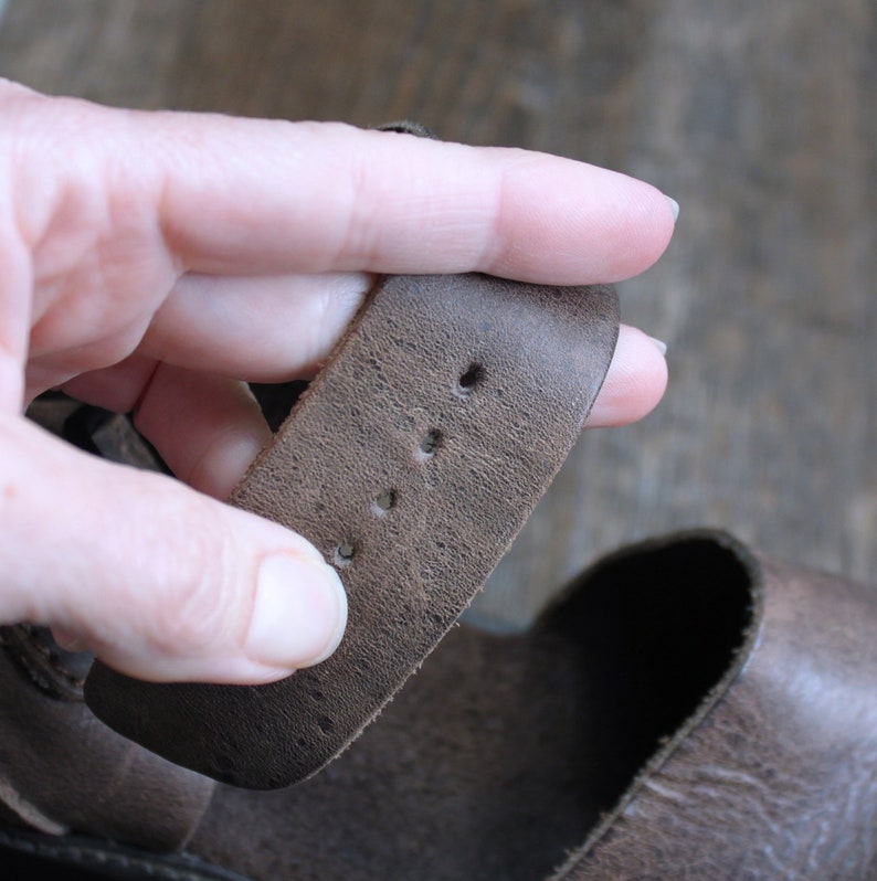 May include: Close-up of a brown leather sandal strap with four holes for adjusting the fit.