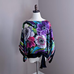 May include: A black silk blouse with a floral print featuring large purple, pink, and white flowers on a black background with green and blue accents. The blouse has a tie at the waist.