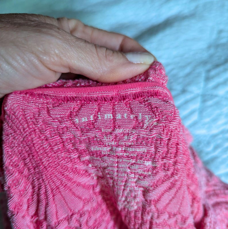 May include: Close-up of a pink, textured fabric with a repeating pattern. The fabric has a scalloped edge and the text "Intimately Free People" is visible. The fabric appears to be a knit or crochet material.