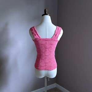 May include: A pink, textured tank top with a square neckline and wide straps. The top is displayed on a mannequin and features a detailed, repeating pattern. The fabric appears to be a knit or crochet material, and the overall style is feminine and vintage-inspired.