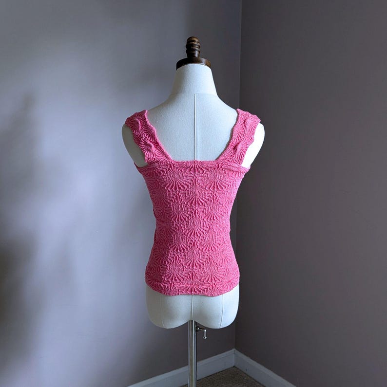 May include: A pink, textured tank top with a square neckline and wide straps. The top is displayed on a mannequin and features a detailed, repeating pattern. The fabric appears to be a knit or crochet material, and the overall style is feminine and vintage-inspired.